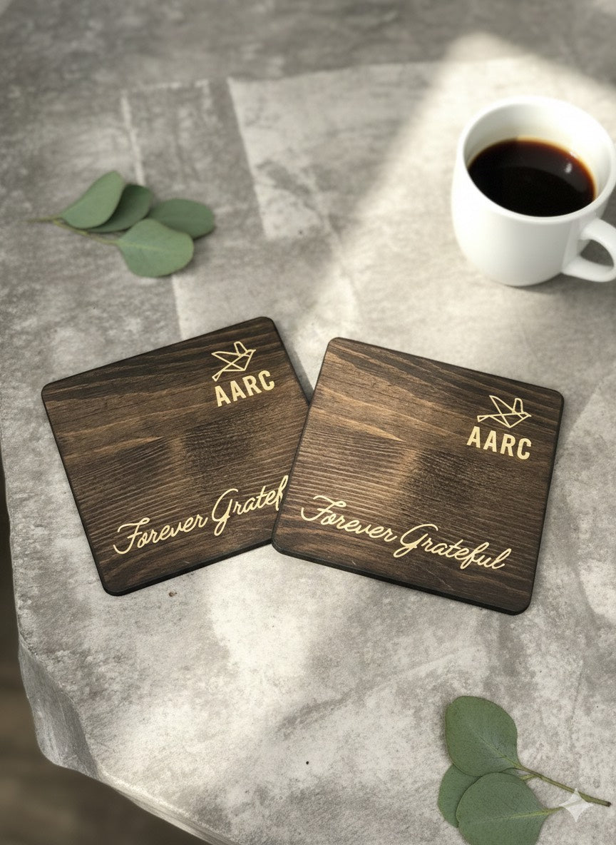 Two square wooden coasters with an engraved AARC logo and Forever Grateful lettering beside a coffee cup.