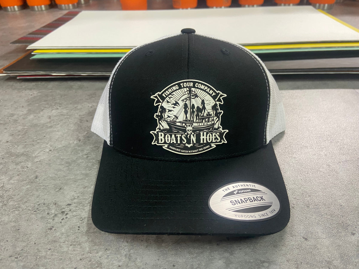 Black and white Boats & Hoes trucker hat with an engraved sailing ship patch, front view.
