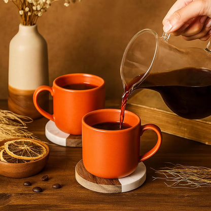 Two orange mugs on round marble and wood coasters while a drink is poured.