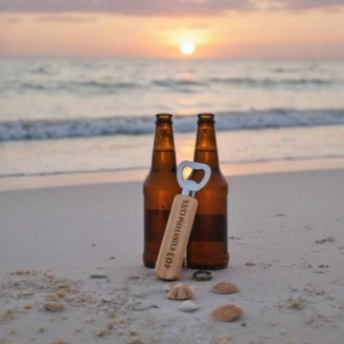 Two beer bottles and a custom engraved bottle opener on a sandy beach with a sunset in the background.