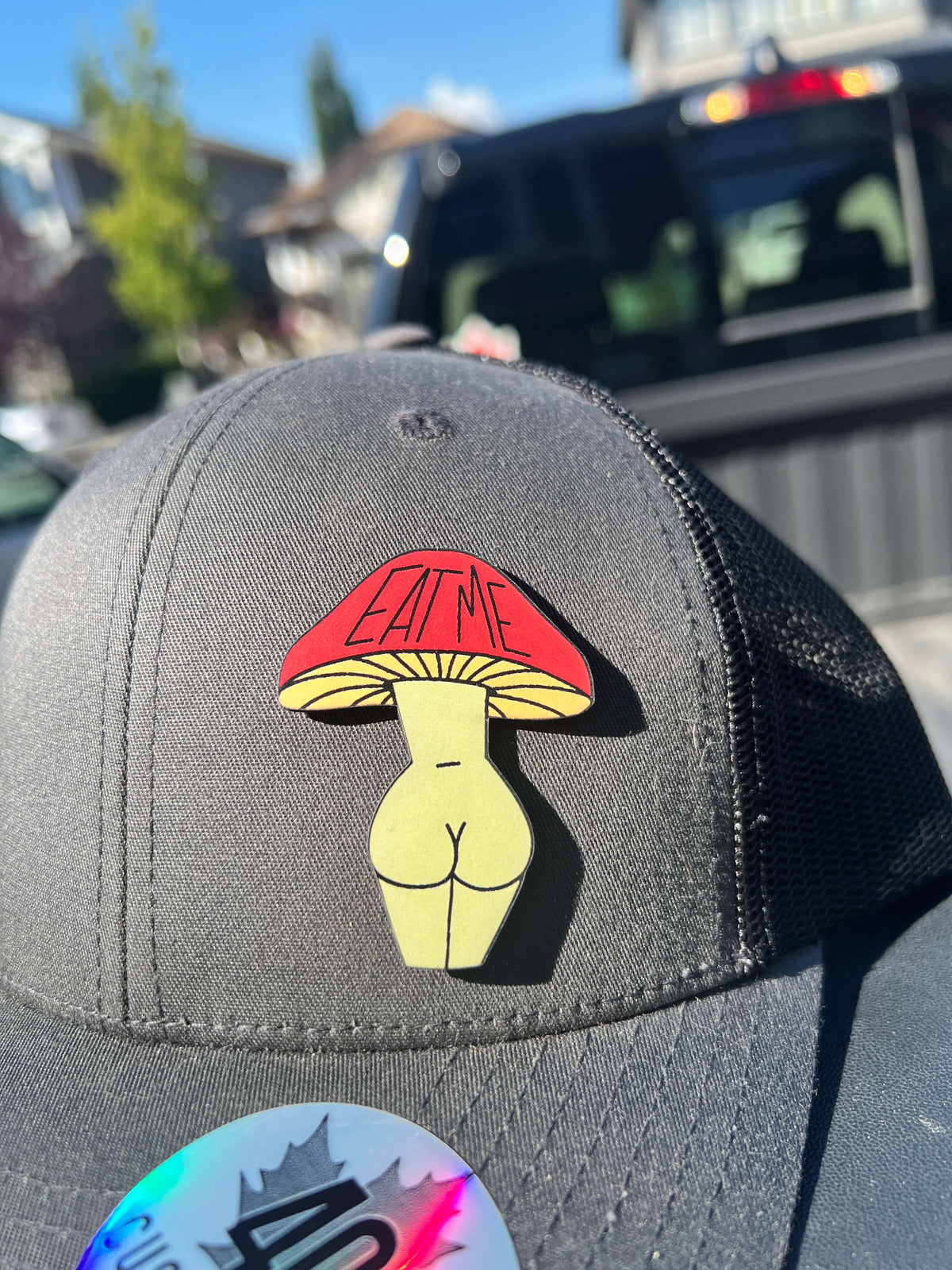 Close-up of the red and yellow Eat Me Mushroom Booty patch on a grey and black trucker hat.