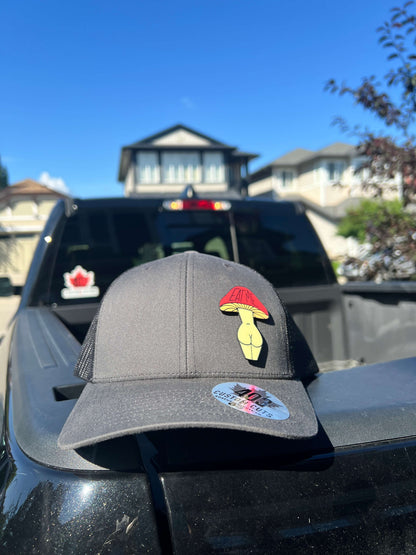 Grey and black Eat Me Mushroom Booty trucker hat with a red and yellow mushroom design, front view outdoors.