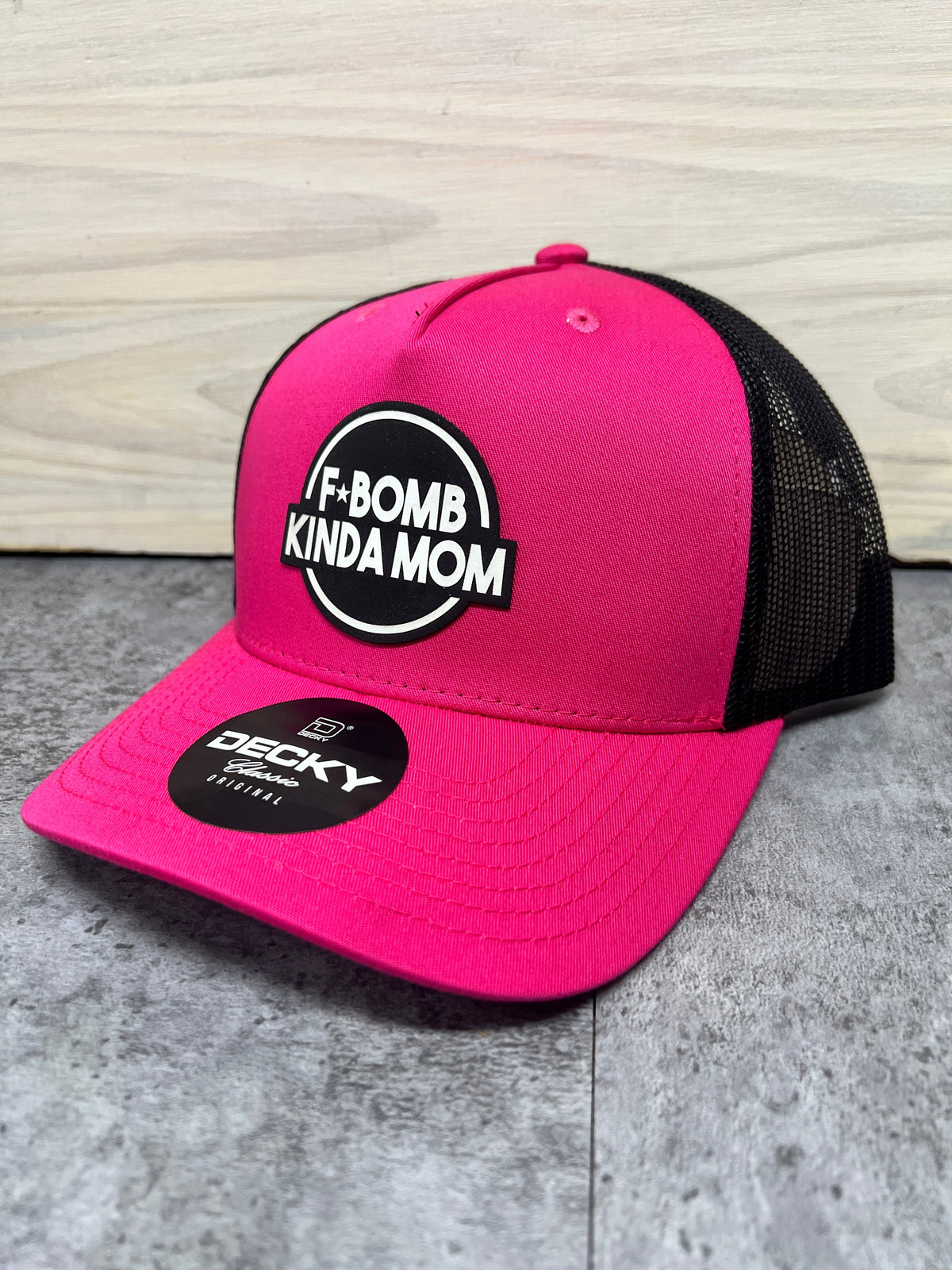 Hot pink and black F*Bomb Kinda Mom trucker hat with a round black and white engraved patch, angled view.