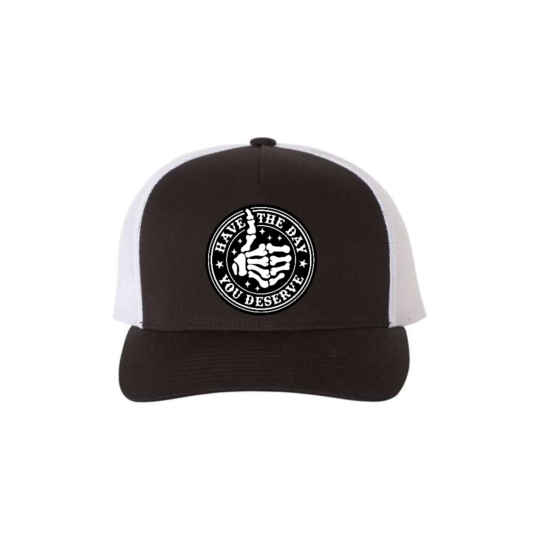 Have the Day You Deserve 5-panel trucker hat in black with white mesh, front product mockup showing a round engraved slogan patch.