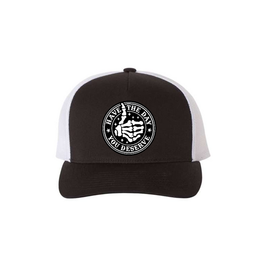 Have the Day You Deserve 5-panel trucker hat in black with white mesh, front product mockup showing a round engraved slogan patch.