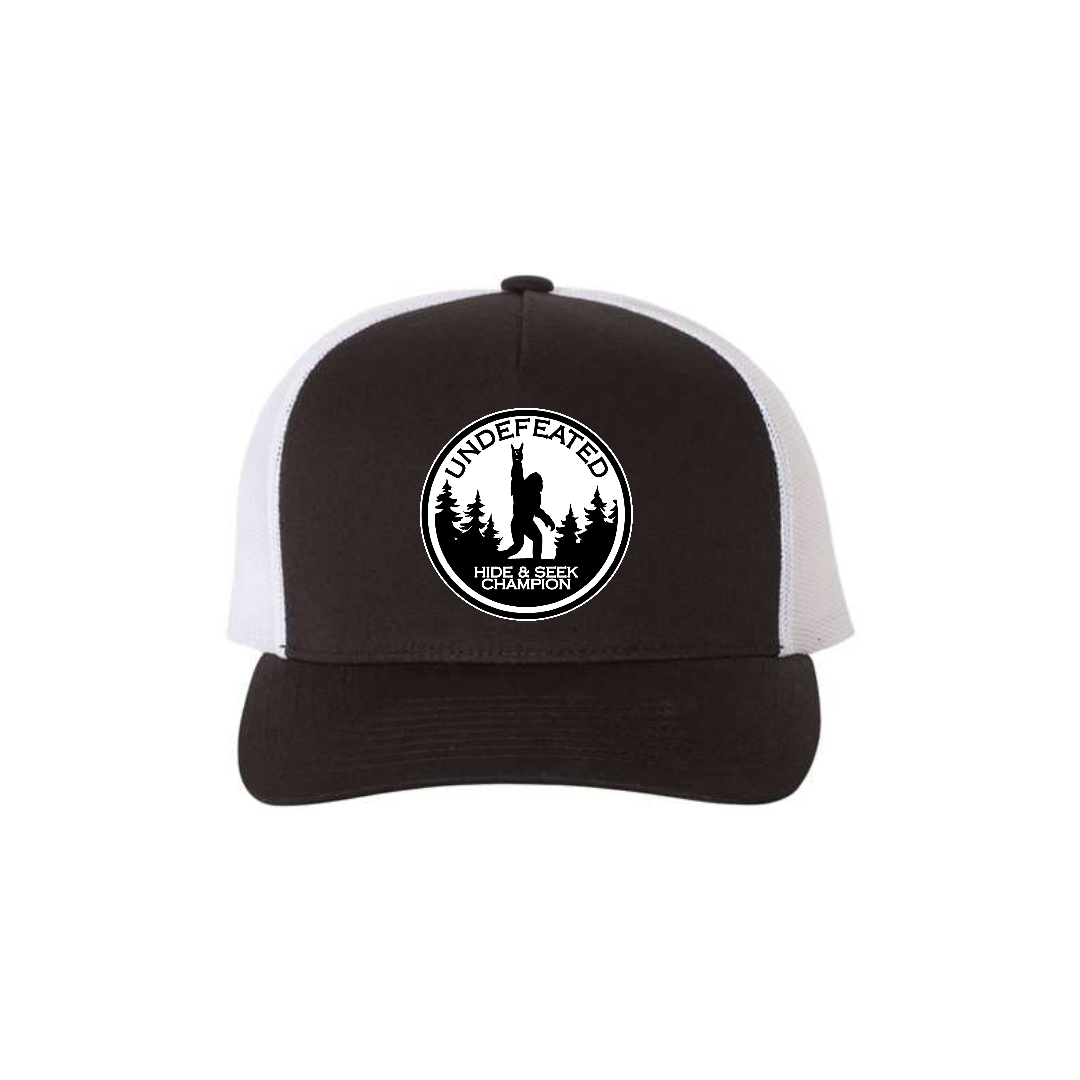 Hide & Seek Champion 5-panel trucker hat in black with white mesh, front product mockup showing a round black and white Bigfoot patch.