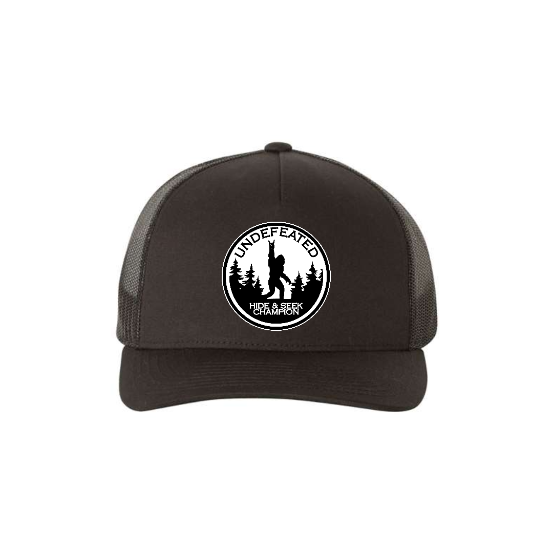 Hide & Seek Champion 5-panel trucker hat in black, front product mockup showing a round black and white Bigfoot patch.