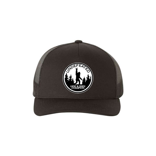 Hide & Seek Champion 5-panel trucker hat in black, front product mockup showing a round black and white Bigfoot patch.
