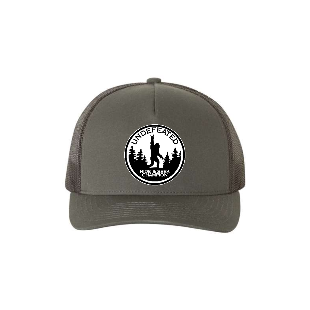 Hide & Seek Champion 5-panel trucker hat in charcoal, front product mockup showing a round black and white Bigfoot patch.