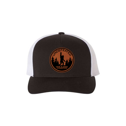 Hide & Seek Champion 5-panel trucker hat in black with white mesh, front product mockup showing a round rawhide Bigfoot patch.