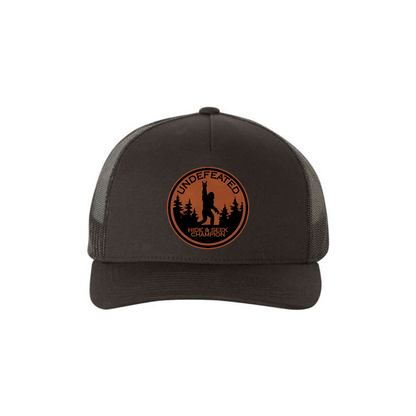 Hide & Seek Champion 5-panel trucker hat in black, front product mockup showing a round rawhide Bigfoot patch.