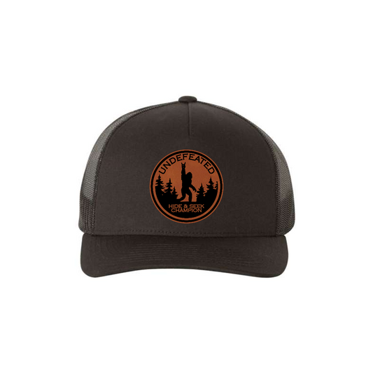 Hide & Seek Champion 5-panel trucker hat in black, front product mockup showing a round rawhide Bigfoot patch.