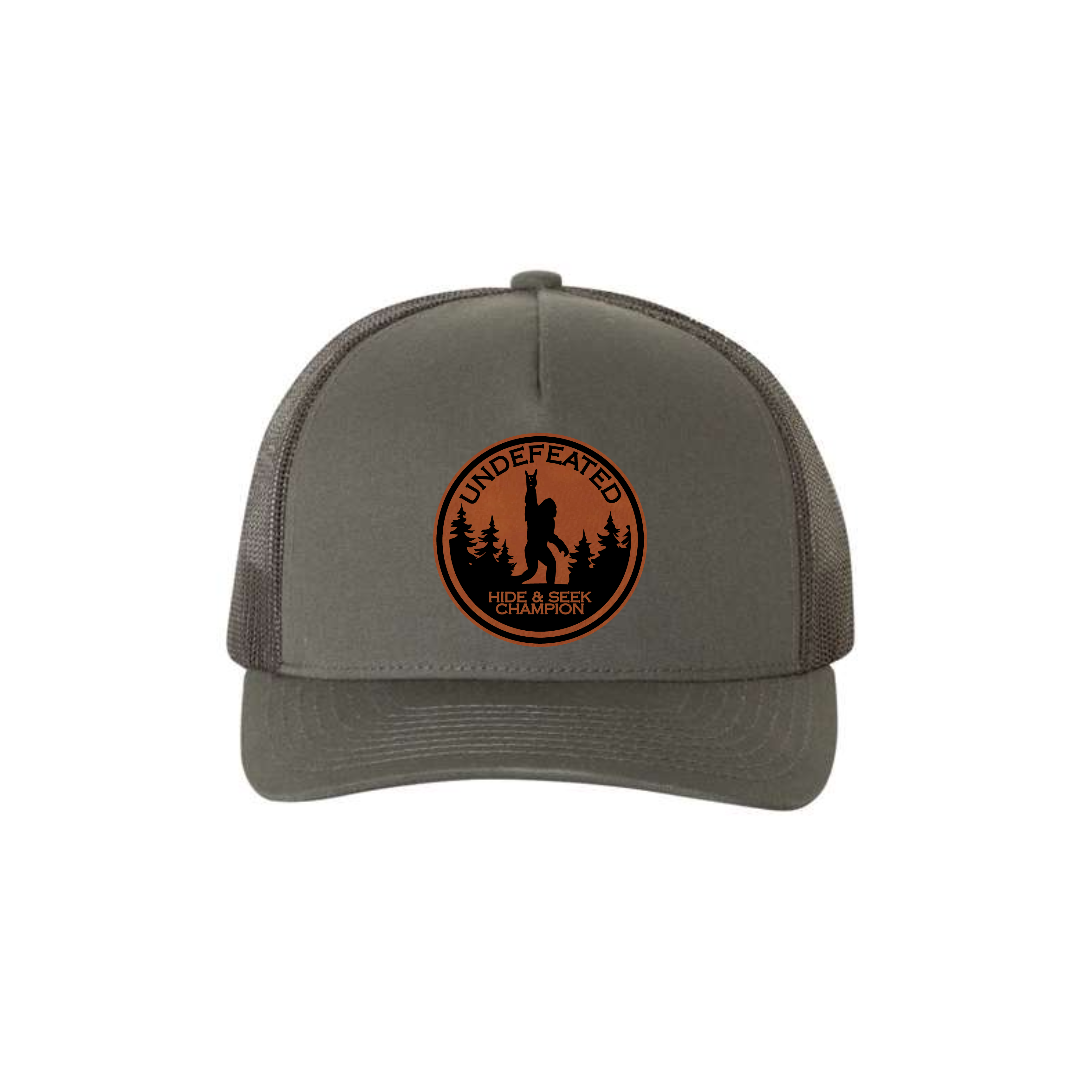 Hide & Seek Champion 5-panel trucker hat in charcoal, front product mockup showing a round rawhide Bigfoot patch.