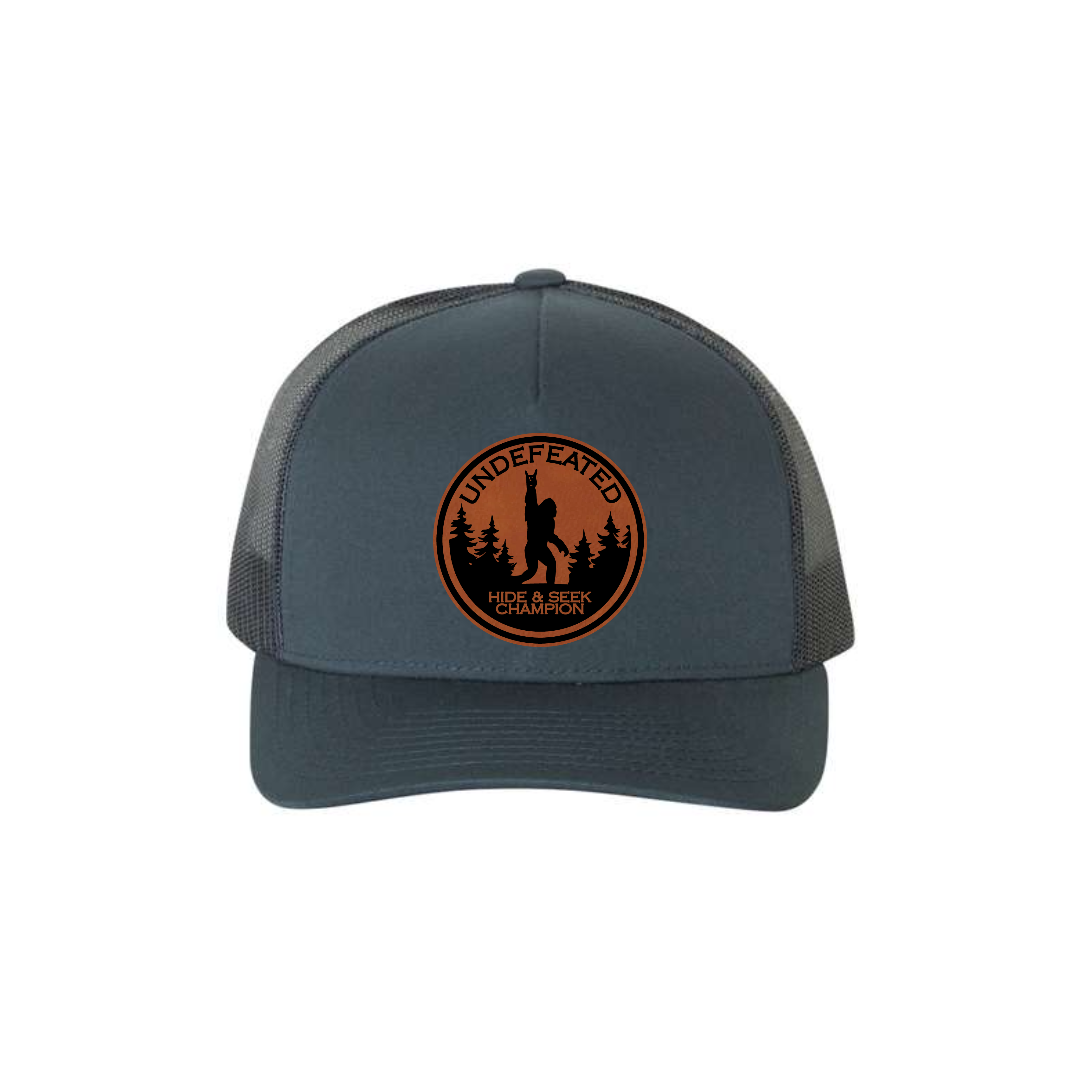 Hide & Seek Champion 5-panel trucker hat in navy, front product mockup showing a round rawhide Bigfoot patch.