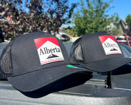 Two black Marly Alberta trucker hats with red and white Alberta Wild Rose Country patches, displayed outdoors.