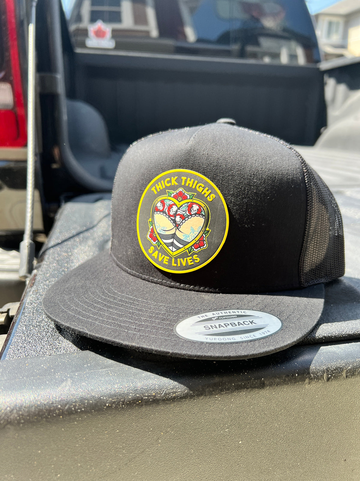 Black Thick Thighs Save Lives trucker hat with a colourful round patch, angled front view on a truck bed.